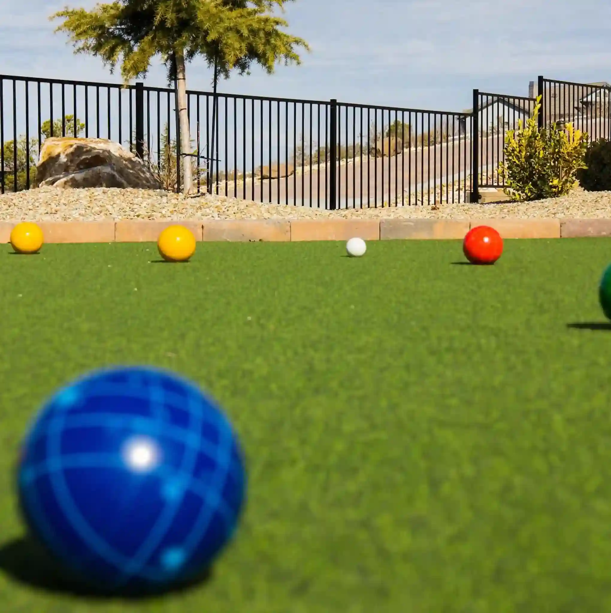 A close-up of colorful bocce balls scattered on vibrant artificial turf in a green bocce court outdoors, with a small tree, rocks, and a black metal fence in the background.