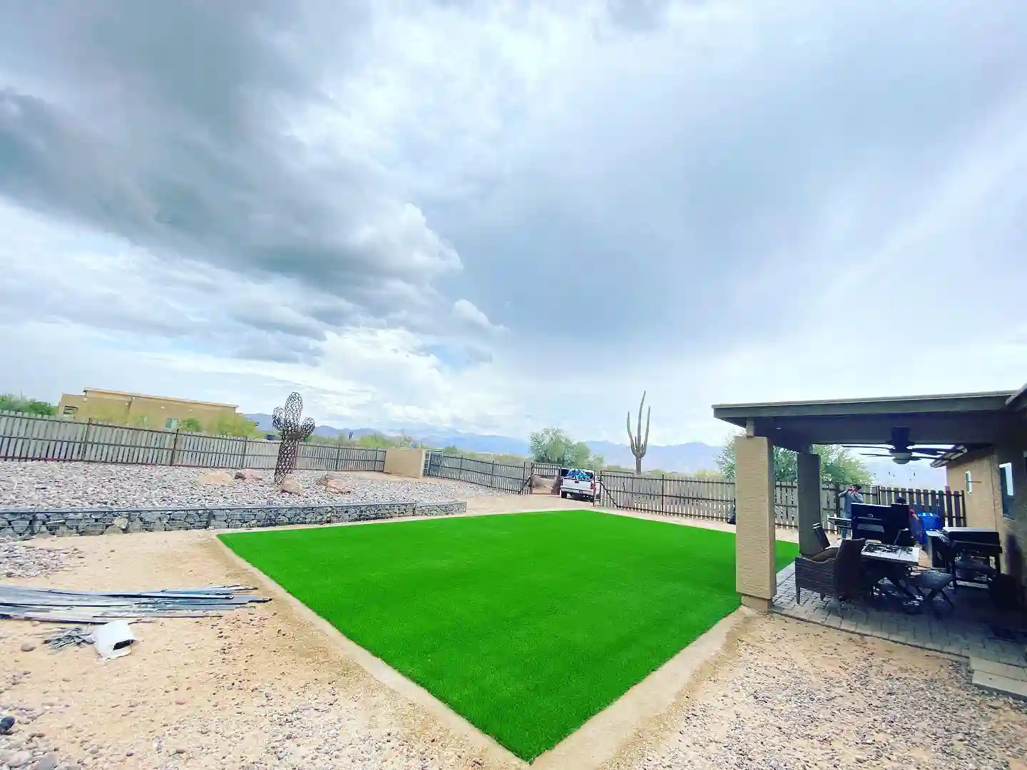 A backyard with bright fake grass bordered by gravel, a covered patio on the right with furniture, cactus sculptures, a wooden fence, and a cloudy sky overhead.