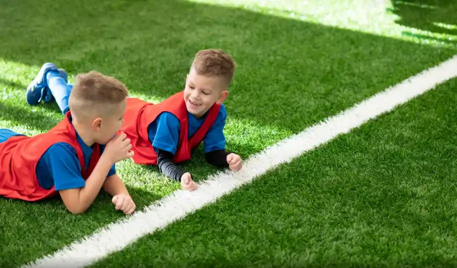 Two young boys wearing blue shirts and red sports vests lie on artificial turf near a white line, smiling and talking to each other.