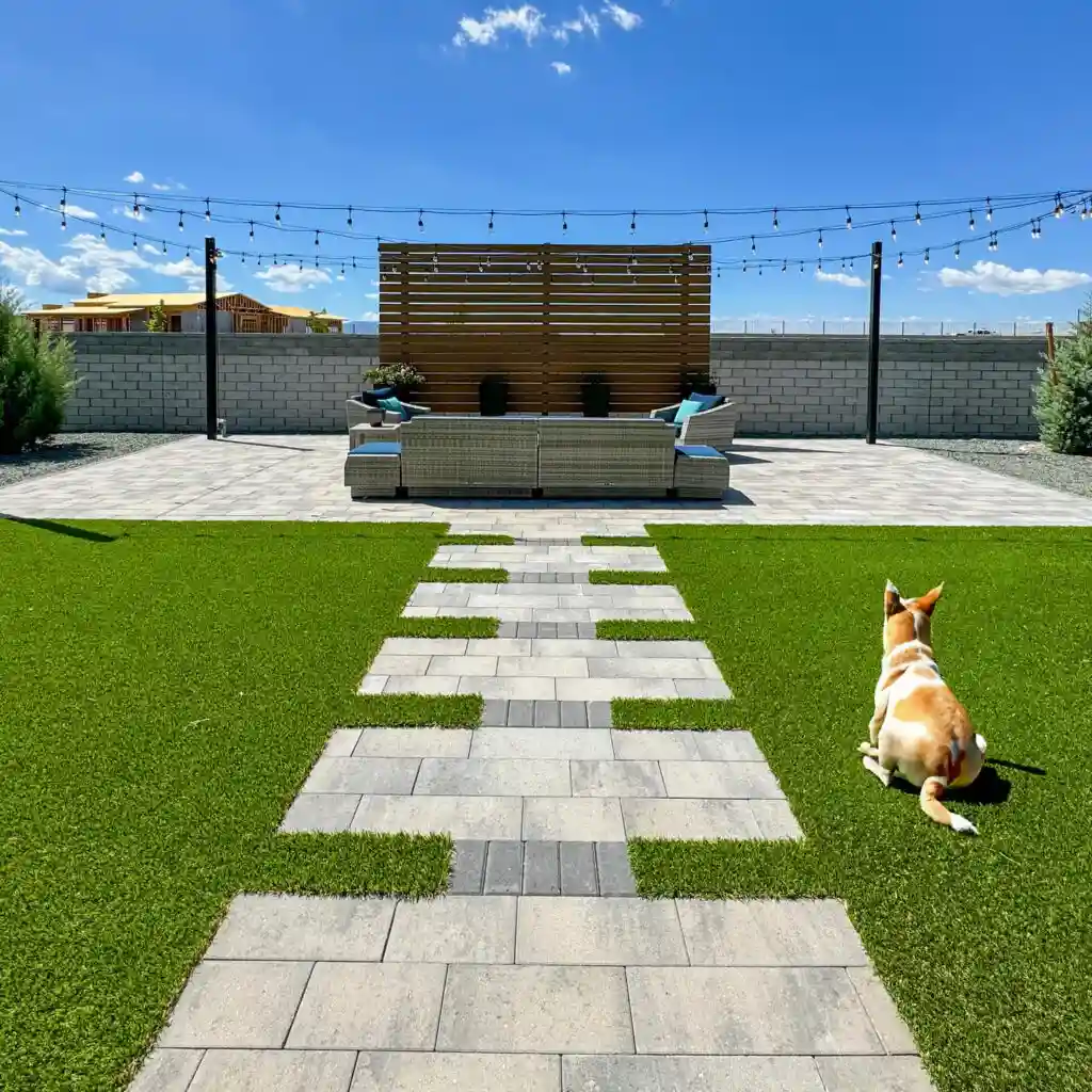 A dog lies on lush Phoenix Synthetic Grass facing a patio with modern seating. Stone pavers create a path, string lights hang above, and the sky is clear—showcasing the benefits of choosing Phoenix Synthetic Grass for your home.