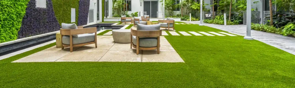 Modern outdoor patio with wooden chairs and round tables on stone tiles, surrounded by lush plants, a vertical garden wall, and artificial green grass that highlights the synthetic turf maintenance benefits you must know in a stylish courtyard setting.