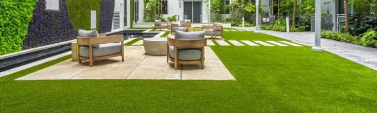 Modern outdoor patio with wooden chairs and round tables on stone tiles, surrounded by lush plants, a vertical garden wall, and artificial green grass that highlights the synthetic turf maintenance benefits you must know in a stylish courtyard setting.