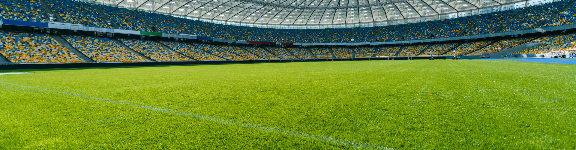 Panoramic view of a large, empty stadium with rows of yellow and blue seats. The field is covered in bright green grass, and a clear sky can be seen through the open roof structure above.