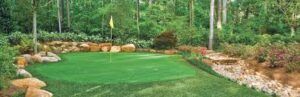 A lush green putting green with a yellow flag is surrounded by trees, rocks, and flowers. A small rocky water feature is visible on the right, nestled among well-manicured grass and bushes.