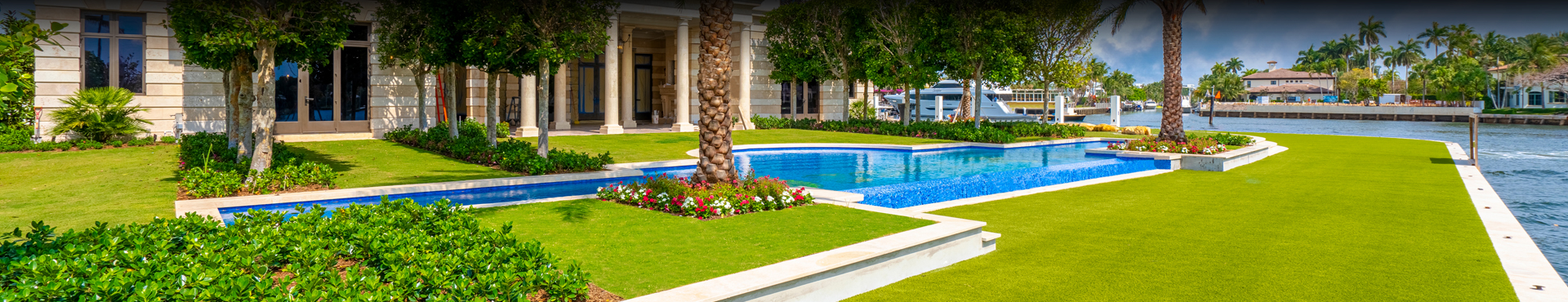 Luxurious waterfront property with manicured lawn, palm trees, and a rectangular swimming pool. The house features a stone exterior with large windows, surrounded by lush greenery, and is adjacent to a waterway with a small dock.