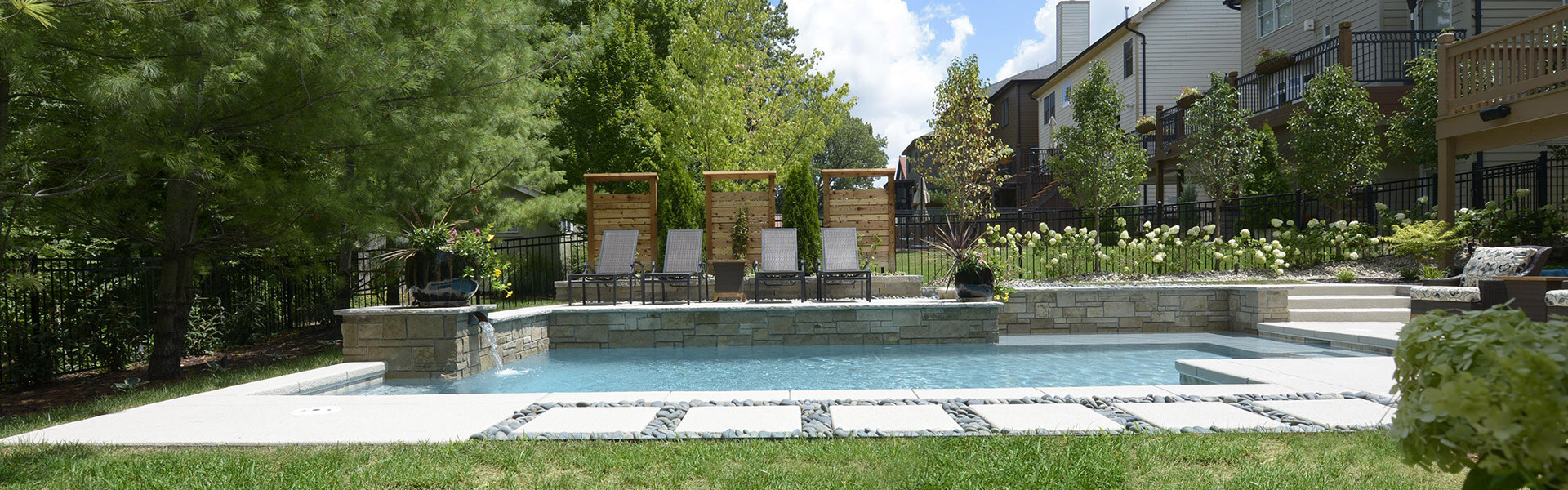 Outdoor pool area with clear blue water, surrounded by stone tiles and grassy edges. Three wooden lounge chairs with cushions are set against a backdrop of trees and shrubs. A row of townhouses is visible in the background on a sunny day.