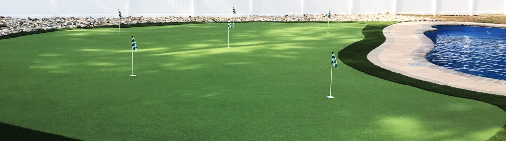A backyard scene featuring a green putting course with several flags, adjacent to a crescent-shaped blue swimming pool. The area is surrounded by a white fence and bordered by rocks, with sunlight creating subtle shadows on the grass.
