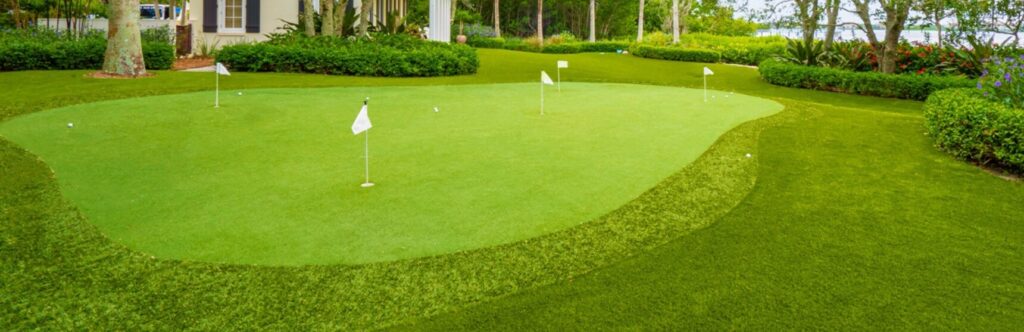 A well-manicured putting green with several white flags on poles, surrounded by neatly trimmed grass and lush greenery. A house with large windows is partially visible on the left, with trees and a body of water in the background.