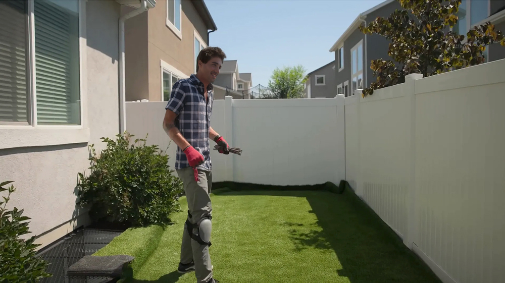 A person wearing gloves and knee pads stands on artificial grass in a narrow backyard, bordered by white fences and houses, showcasing a sunny day of DIY artificial grass installation.
