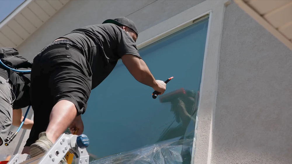 A person on a ladder uses a squeegee to clean or apply film to a large exterior window on a light-colored house—an easy guide for those considering DIY projects like artificial grass installation. The individual wears black clothing and a backward cap.