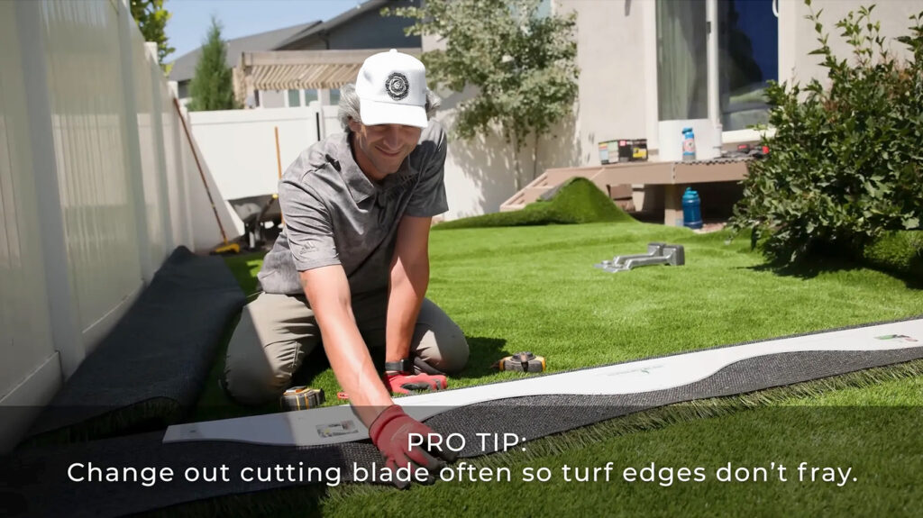 A person kneels on artificial turf, using a cutting tool to trim a roll during DIY artificial grass installation. The scene is outdoors in a backyard. Text reads: “PRO TIP: Change out cutting blade often so turf edges don’t fray.”.